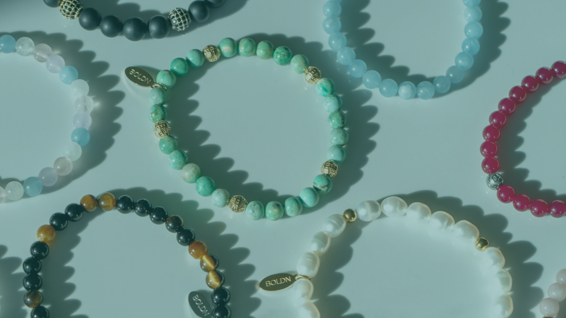 A collection of BOLDN beaded bracelets displayed on a reflective white surface, featuring various gemstones such as turquoise, aquamarine, tiger’s eye, and freshwater pearls, with gold and silver accents