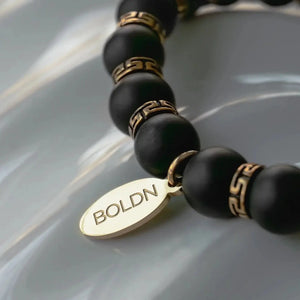 Close-up of a black beaded bracelet with gold Greek key-patterned spacers and a gold BOLDN tag, placed on a reflective surface