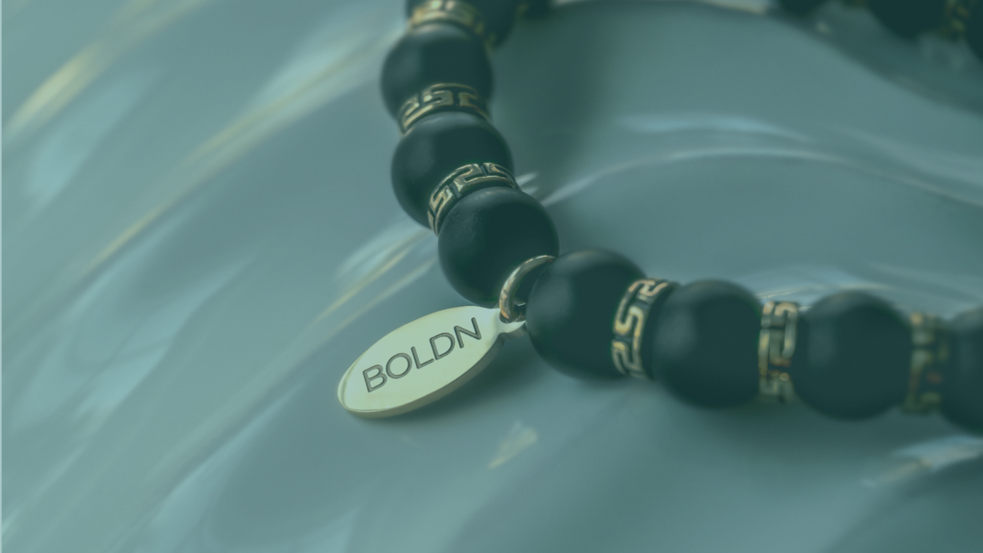 Close-up of a black beaded bracelet with gold Greek key-patterned spacers and a gold BOLDN tag, placed on a reflective surface